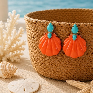 Puede incluir: Un par de pendientes colgantes con colgantes de concha en forma de abanico de color naranja brillante, suspendidos de pequeñas piedras turquesa en forma de lágrima. Los pendientes están sujetos al borde de una cesta tejida de color tostado, mostrando un contraste vibrante entre las conchas naranjas y las piedras azules.