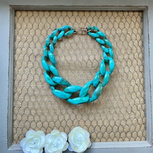 May include: A turquoise blue chunky chain necklace with a gold clasp. The necklace is displayed on a brown burlap background.