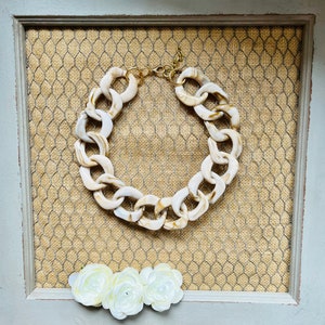 May include: A chunky white and brown resin chain necklace with a gold clasp. The necklace is displayed on a brown woven background.