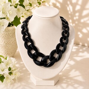 May include: A chunky statement necklace featuring large, glossy black oval links connected in a continuous chain design. The necklace is displayed on a smooth, off-white bust, highlighting the bold, reflective surface of the dark plastic material.