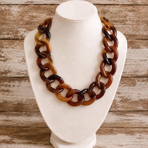 May include: A chunky chain link necklace featuring large, interlocking oval links in a translucent, marbled brown and amber tortoiseshell pattern. The necklace is displayed on a smooth, off-white, neck-shaped jewellery bust stand.