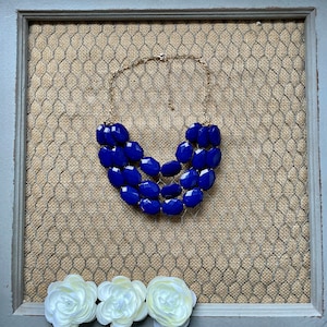 Peut inclure: Un collier à trois rangs composé de grosses perles facettées bleu foncé. Le collier est sur une chaîne dorée et est présenté sur un fond en toile de jute brune.