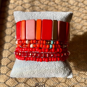 Op de afbeelding: Een stapel rode, oranje en roze kralenarmbanden met gouden accenten. De armbanden liggen op een grijs fluwelen kussen. Het woord "Rood" staat onder het kussen.
