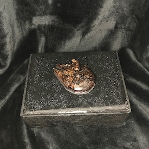Puede incluir: Una caja negra con un emblema metálico de bronce del Halcón Milenario en la tapa. La caja tiene un diseño brillante y estrellado en la parte superior y los lados, sobre un fondo oscuro.