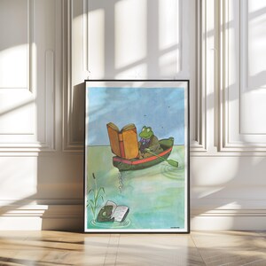 May include: A framed art print featuring a whimsical illustration of a frog in a boat reading a book. The frog is wearing a jacket and a bow tie. Another frog is reading a book in the water. The background is a blue sky and water.