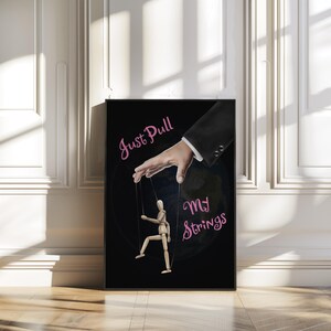 May include: A framed art print on a black background. The image features a hand in a suit sleeve pulling strings attached to a wooden puppet. The text "Just Pull My Strings" is in pink cursive.