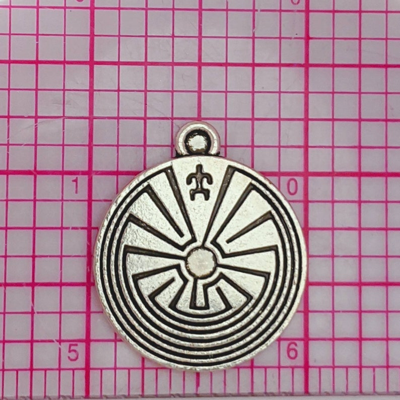 May include: Silver charm with a circular design featuring a labyrinth pattern and a symbol in the center. The charm has a loop at the top for attaching to a necklace or bracelet.