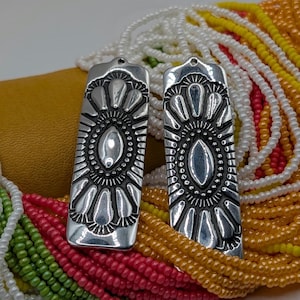 May include: Two silver-toned rectangular pendants with an oval center and floral design. The pendants are displayed against a backdrop of colorful beaded necklaces in red, green, yellow, and white. The pendants are likely for jewelry making.