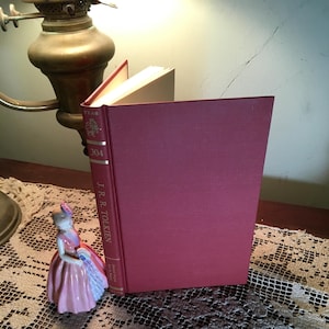 May include: A hardcover book by J.R.R. Tolkien with a burgundy cover, standing upright on a table. The book's spine displays the author's name and the number 304. A small porcelain figurine in a pink dress is next to the book.
