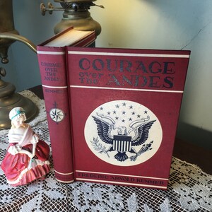 May include: A vintage book titled "Courage Over The Andes" with a red cover and a white emblem featuring an eagle and stars. The book is displayed on a lace tablecloth, with a porcelain figurine nearby.