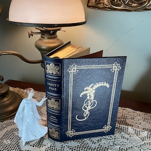 May include: A hardcover book titled "Vanity Fair" with gold lettering and decorative accents. The book is dark blue with a gold design on the cover. A white porcelain figurine stands nearby, and a vintage lamp is in the background.