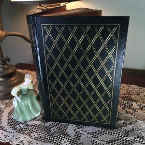May include: A hardcover book with a black cover and a gold diamond pattern. The book is open, revealing the pages inside. A porcelain figurine in a green dress is next to the book. A vintage lamp is in the background.