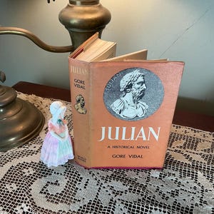 Puede incluir: Un libro abierto titulado "JULIAN" de Gore Vidal, con un retrato de un hombre en la portada. Una peque&ntilde;a figura de porcelana de una mujer con un vestido est&aacute; a la izquierda del libro. El libro est&aacute; sobre un mantel de encaje.