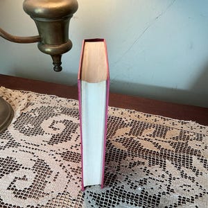 Puede incluir: Un libro de tapa dura con una cubierta y lomo rosas, de pie. Las p&aacute;ginas son blancas y el borde superior de las p&aacute;ginas es visible. El libro est&aacute; sobre una superficie de madera, con una l&aacute;mpara de lat&oacute;n en el fondo.