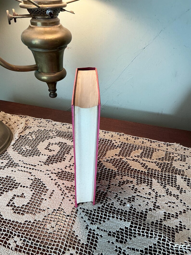 Puede incluir: Un libro de tapa dura con una cubierta y lomo rosas, de pie. Las p&aacute;ginas son blancas y el borde superior de las p&aacute;ginas es visible. El libro est&aacute; sobre una superficie de madera, con una l&aacute;mpara de lat&oacute;n en el fondo.