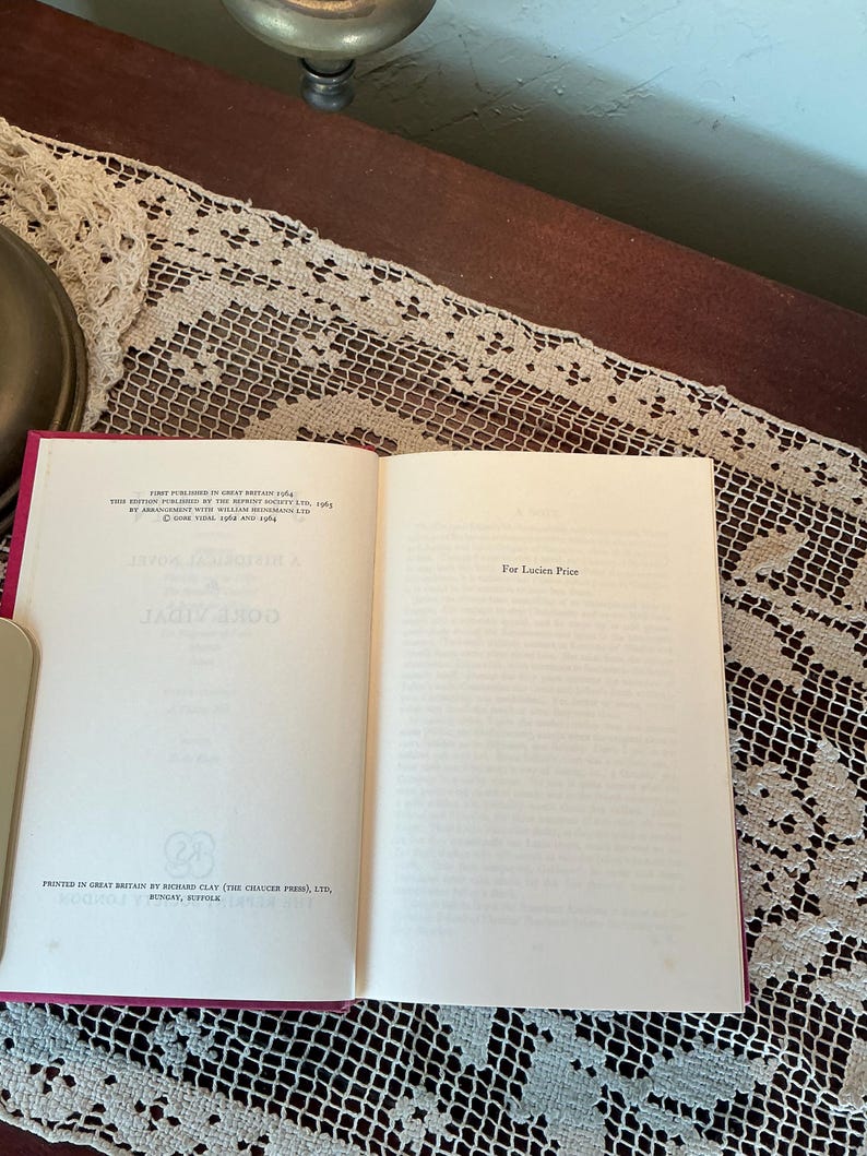 Puede incluir: Un libro abierto con una cubierta roja y p&aacute;ginas blancas. El texto de la p&aacute;gina izquierda dice "GORE VIDAL" y la p&aacute;gina derecha dice "For Lucien Price". El libro est&aacute; sobre una mesa con un camino de mesa de encaje.
