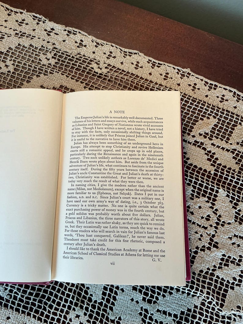 Puede incluir: Libro abierto con texto sobre una mesa con un mantel de encaje blanco. El texto incluye el t&iacute;tulo "A NOTE" y trata sobre la vida del emperador Juliano. Las p&aacute;ginas del libro son de color crema con texto negro.