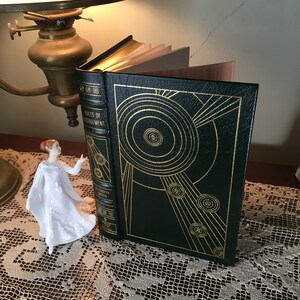 May include: A hardcover book with a dark green cover and gold geometric designs. The book is open, revealing the pages. The title "Rules of Engagement" is visible on the spine. A small white figurine is next to the book.