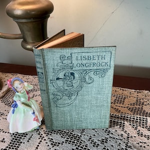 May include: A vintage book titled "Lisbeth Longfrock" with a teal cover and ornate design. The book is open, revealing aged pages. A porcelain figurine of a woman in a green dress and a blue bow is next to the book. The scene is lit by a brass lamp.