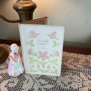 May include: A vintage book titled "Friendly Counsels by F.B. Meyer" with a floral design on the cover. A small porcelain figurine of a woman in a bonnet and dress is next to the book. The book and figurine are on a lace tablecloth.