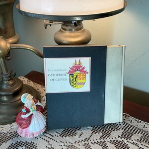 May include: A hardcover book titled "The Hours of Catherine of Cleves" with a decorative label featuring a coat of arms. The book has a dark blue cover and a silver spine. A porcelain figurine is in the foreground.