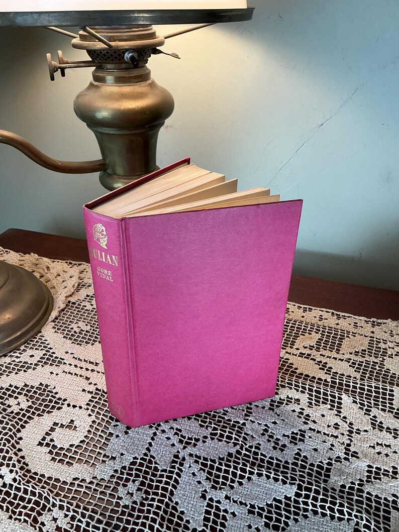 Puede incluir: Un libro de tapa dura rosa brillante, titulado "Julian Gore Vidal". El libro est&aacute; ligeramente abierto, revelando las p&aacute;ginas amarillentas. El libro est&aacute; sobre una mesa con un mantel de encaje, con una l&aacute;mpara de lat&oacute;n en el fondo.