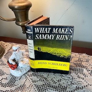 May include: A vintage copy of "What Makes Sammy Run?" by Budd Schulberg. The book has a black cover with yellow text and a landscape image. A small porcelain figurine and a brass lamp are also visible.