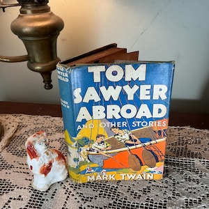 May include: A vintage copy of "Tom Sawyer Abroad and Other Stories" by Mark Twain. The book has a blue cover with bold white and yellow lettering and a colorful illustration. A small porcelain dog figurine sits nearby.