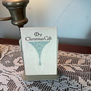 May include: A vintage Christmas card titled "My Christmas Gift" in a light green holder. The card features an ornate teal design and text. The scene is set on a table with a lace doily and a brass lamp.