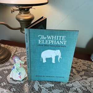May include: A teal book titled "The White Elephant And Other Tales from India" with a white elephant illustration. The book is propped up on a wooden surface next to a porcelain figurine and a vintage lamp.