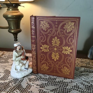 May include: A maroon hardcover book titled "The Prisoner of Zenda" with gold floral designs. The book is next to a porcelain figurine of a woman playing a stringed instrument. The book is on a lace tablecloth.