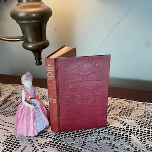 May include: A vintage book with a red cover and embossed design, titled "The Phantom 'Rickshaw and City of Dreadful Night" by Rudyard Kipling. A porcelain figurine in a pink dress and a brass lamp are also visible.