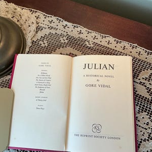 Puede incluir: Un libro abierto titulado "JULIAN" de Gore Vidal. El libro es una novela hist&oacute;rica, publicada por The Reprint Society London. El libro tiene un lomo rojo y est&aacute; sobre un mantel de encaje.