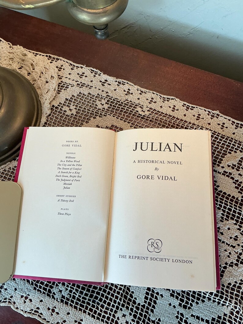 Puede incluir: Un libro abierto titulado "JULIAN" de Gore Vidal. El libro es una novela hist&oacute;rica, publicada por The Reprint Society London. El libro tiene un lomo rojo y est&aacute; sobre un mantel de encaje.