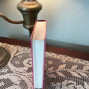 Puede incluir: Un libro de tapa dura con una cubierta rosa y p&aacute;ginas blancas. El libro est&aacute; de pie sobre una mesa con un tapete de encaje. Una l&aacute;mpara de lat&oacute;n vintage est&aacute; en el fondo.