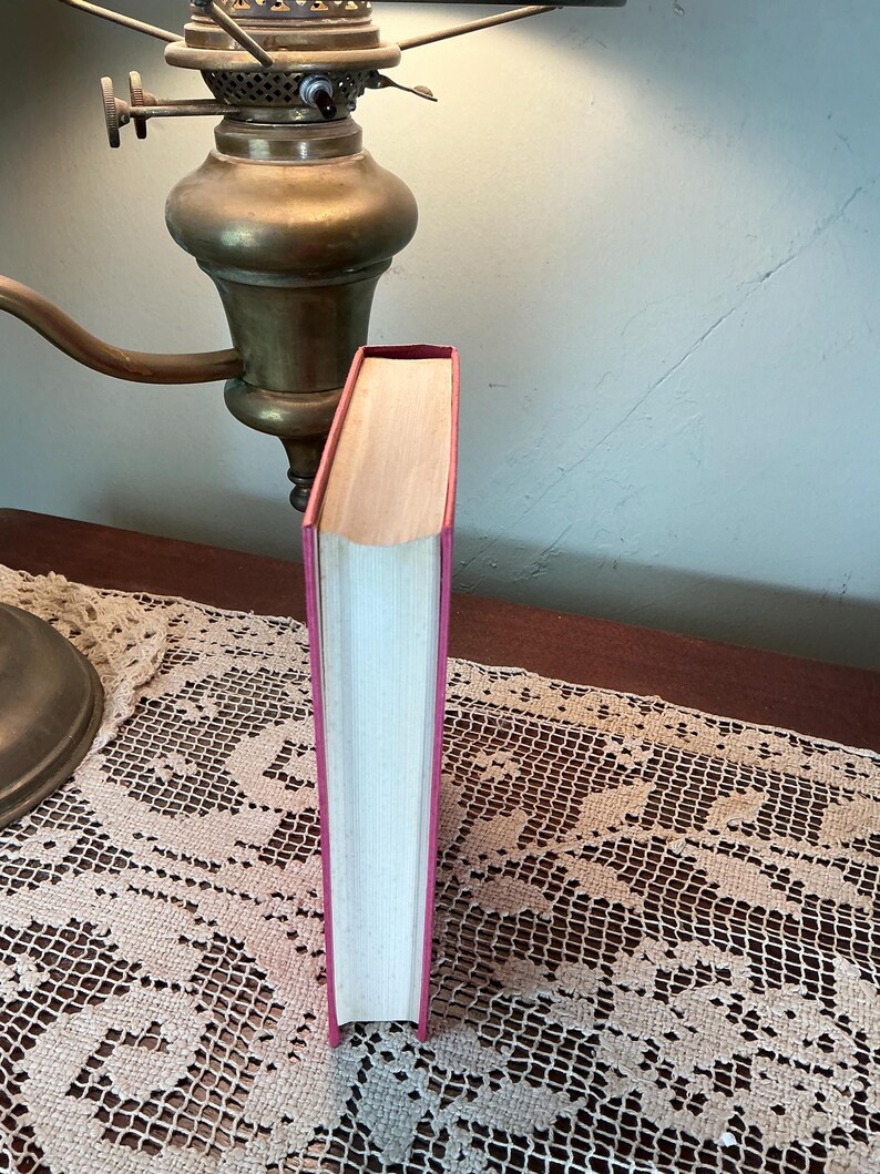 Puede incluir: Un libro de tapa dura con una cubierta rosa y p&aacute;ginas blancas. El libro est&aacute; de pie sobre una mesa con un tapete de encaje. Una l&aacute;mpara de lat&oacute;n vintage est&aacute; en el fondo.