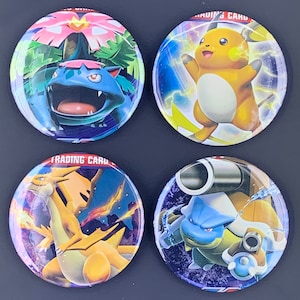 Puede incluir: Cuatro botones redondos del Juego de Cartas Coleccionables Pokémon. Los botones presentan diferentes personajes Pokémon, incluyendo Venusaur, Pikachu, Charizard y Blastoise. Los botones son azules, amarillos, rojos y morados.