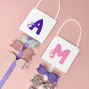 May include: Two white square plaques with the letters "A" and "M" in purple and pink, and heart accents. Each plaque has a white handle. Below are various coloured hair bows attached to ribbons.