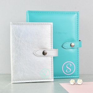 May include: Two earring storage boxes, one silver and one turquoise. The silver box has the text "The Little Book of Earrings" and the turquoise box has the text "Book of Earrings". Both boxes have a letter initial on the front, "R" on the silver box and "S" on the turquoise box.  A pair of white earrings are on a pink surface.