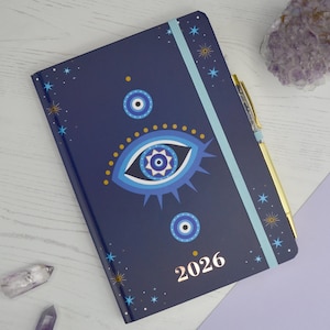 Op de afbeelding: Een marineblauwe 2026-planner met een oogontwerp en gouden accenten. De cover heeft een groot blauw oogsymbool, kleinere oogsymbolen en sterpatronen. Een gouden pen rust op de planner en een lichtblauwe elastische band houdt hem gesloten.