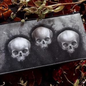 Puede incluir: Un estuche negro con cremallera que presenta una ilustración en escala de grises de tres cráneos humanos. Los cráneos están centrados en el estuche, con un fondo oscuro y sutiles detalles en forma de estrella. El estuche es rectangular y parece ser de tela.