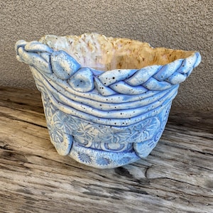 May include: A handcrafted ceramic bowl with a light blue exterior and a textured, braided rim. The bowl features embossed floral patterns and a speckled finish. The interior has a tan and cream color. The bowl sits on a weathered wooden surface.