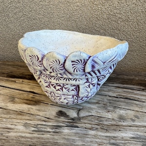 May include: A handcrafted ceramic bowl with a speckled white interior and a detailed exterior. The bowl features a light purple and white color scheme with intricate geometric and floral patterns. The rim has a scalloped design.