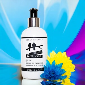 May include: A white bottle of organic hand wash with a black and white label. The label says "Vegan Friendly", "Organic Hand Wash", "No 56", "Burds of Paradise", "Handmade in Scotland", and "Twa Burds". The bottle is sitting on a blue and purple background with yellow and blue flowers.