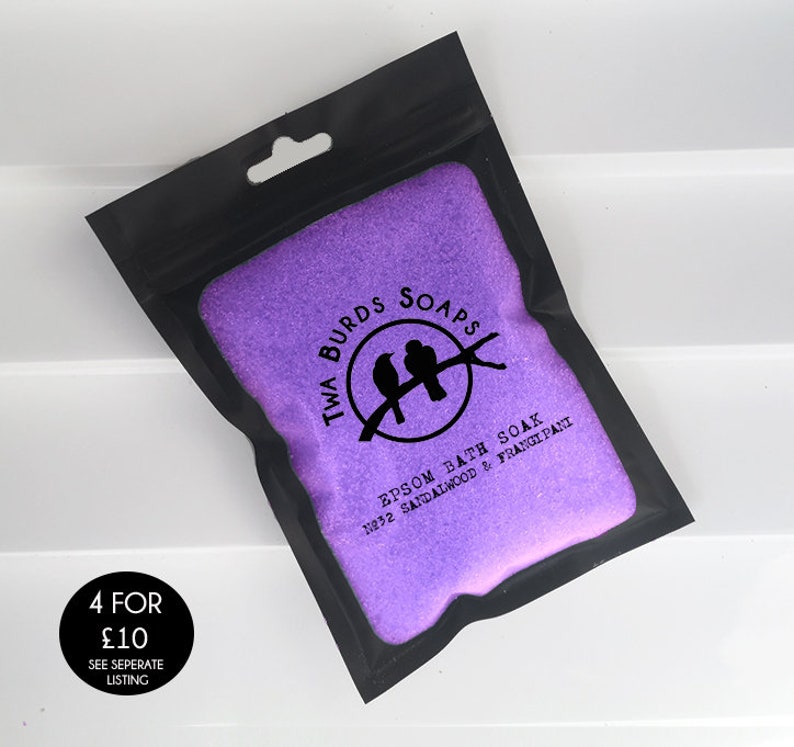 May include: A black resealable pouch filled with purple bath salts. The pouch has a white label with the text "Twa Burds Soaps" and a logo of two birds on a branch. The label also includes the text "Epsom Bath Soak 1932 Sandalwood & Frangipani".