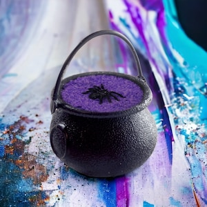 May include: A black cauldron with a purple, glittery interior and a black spider on top. The cauldron is sitting on a colourful, abstract background.