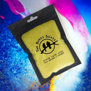May include: A black pouch with a yellow zipper contains a yellow bath soak. The pouch has a logo that says "Twa Burds Soaps" with two birds on a branch. The text on the pouch reads "Epsom Bath Soak No.41 Rhubarb & Custard."