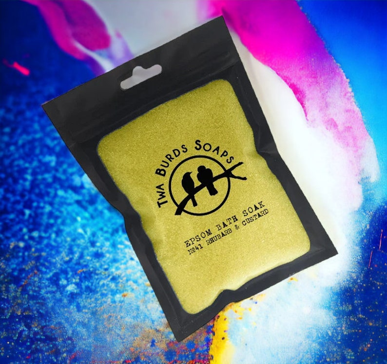 May include: A black pouch with a yellow zipper contains a yellow bath soak. The pouch has a logo that says "Twa Burds Soaps" with two birds on a branch. The text on the pouch reads "Epsom Bath Soak No.41 Rhubarb & Custard."