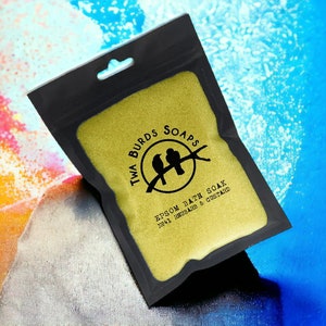 May include: A black pouch with a gold zipper contains a yellow bath soak. The pouch has a white label with the text "Twa Burds Soaps" and a logo of two birds on a branch. The text "Epsom Bath Soak No.41 Rhubarb & Custard" is printed on the label.