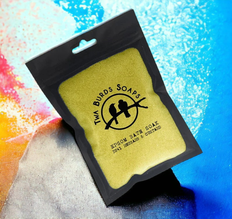 May include: A black pouch with a gold zipper contains a yellow bath soak. The pouch has a white label with the text "Twa Burds Soaps" and a logo of two birds on a branch. The text "Epsom Bath Soak No.41 Rhubarb & Custard" is printed on the label.
